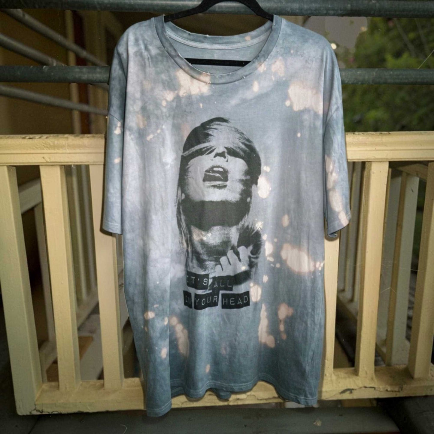 A light blue-gray acid-wash graphic t-shirt hanging on a black hanger against a weathered wooden railing. The shirt features a black-and-white graphic of a blindfolded woman with the text 'IT'S ALL IN YOUR HEAD' printed on the front.