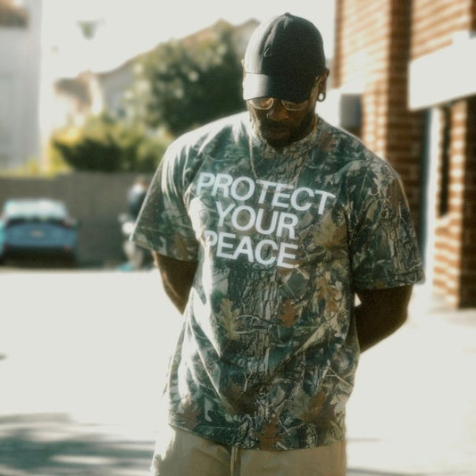 Man wearing a camouflage t-shirt with 'PROTECT YOUR PEACE' text outdoors.