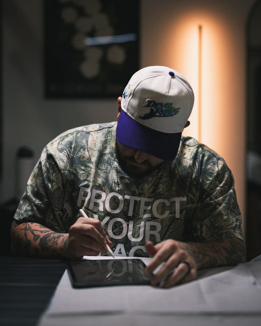 Man wearing a cap and camouflage shirt with text, sitting at a table with a tablet.