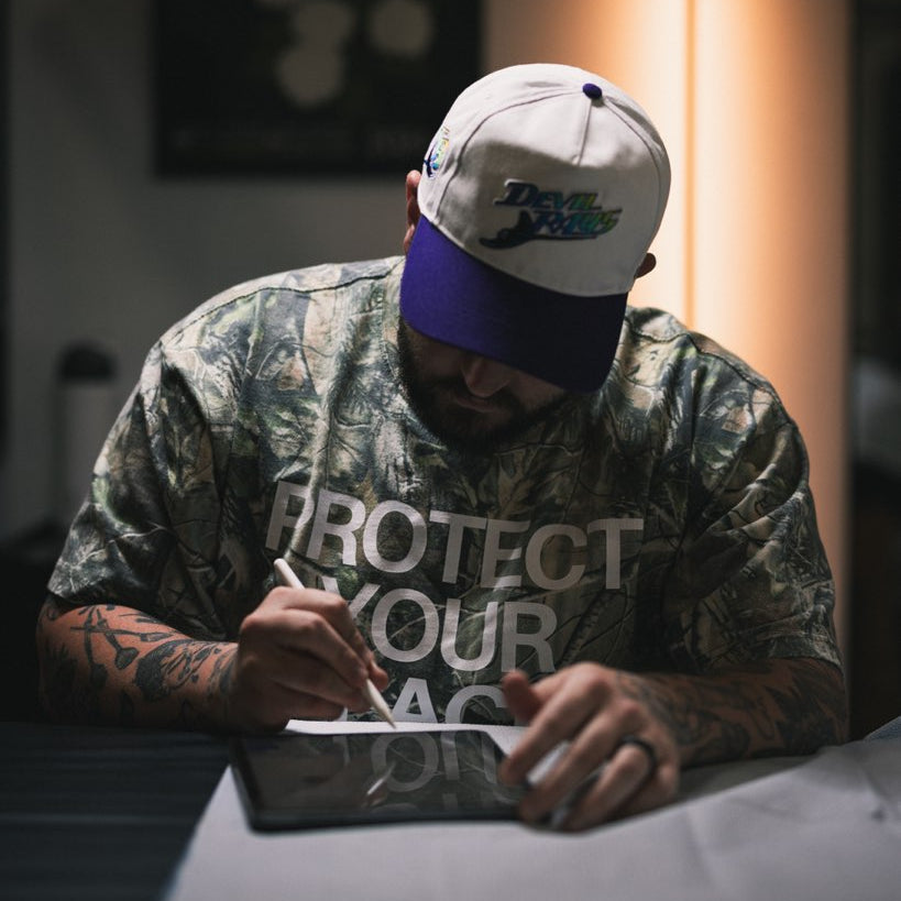 Man wearing a cap and camouflage shirt with text, sitting at a table with a tablet.