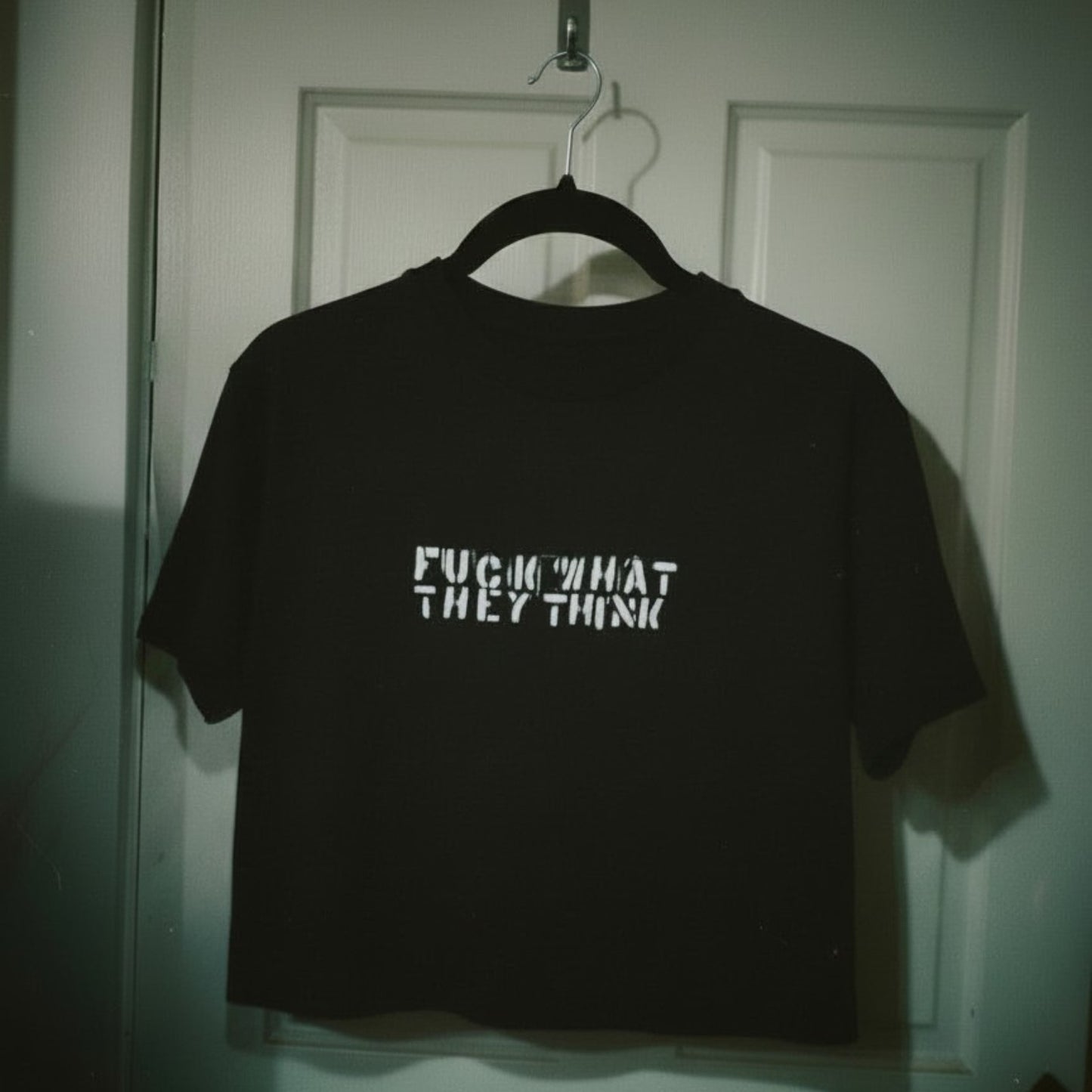 A black short-sleeve t-shirt hanging on a black hanger against a white paneled door. The shirt features the text 'FUCK WHAT THEY THINK' printed in a white, distressed stencil font across the chest.