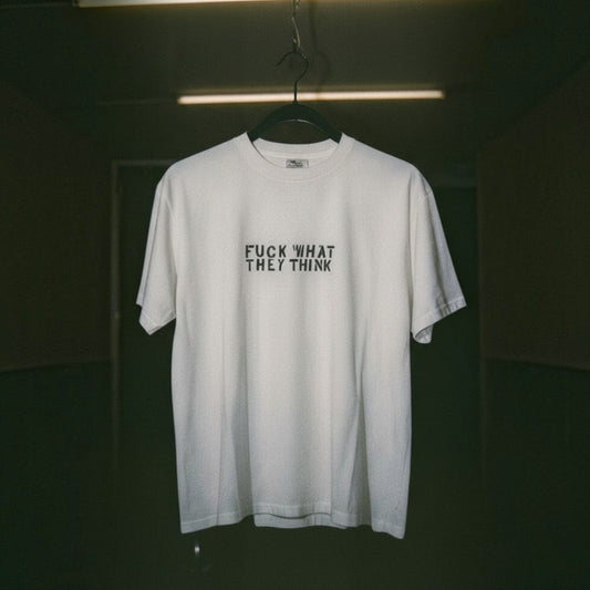 A white short-sleeve t-shirt hanging on a black hanger against a dark, moody background. The shirt features the text 'FUCK WHAT THEY THINK' printed in black capital letters across the chest.