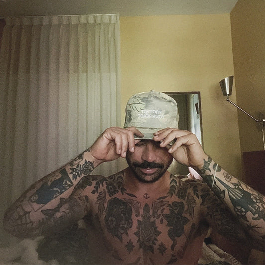 Tattooed man wearing a camo snapback hat embroidered with “Protect Your Peace” by Comfortable Silence, lifestyle photo indoors