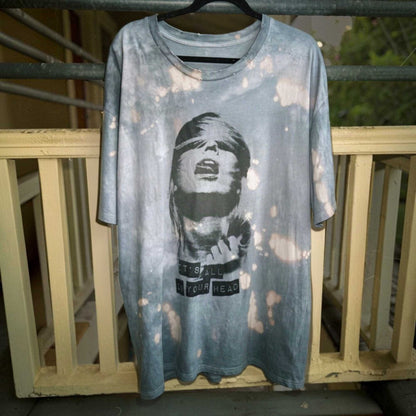 A light blue-gray acid-wash graphic t-shirt hanging on a black hanger against a weathered wooden railing. The shirt features a black-and-white graphic of a blindfolded woman with the text 'IT'S ALL IN YOUR HEAD' printed on the front.
