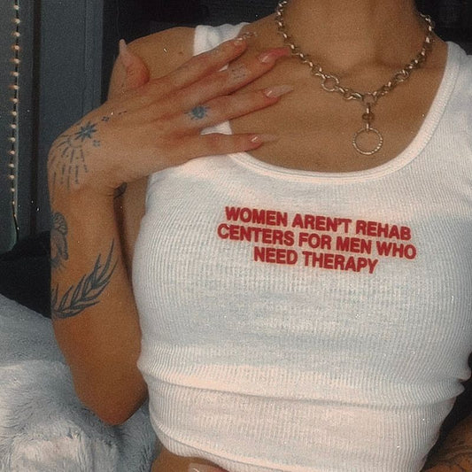 A woman wearing a white ribbed tank top with red text that reads "Women Aren't Rehab Centers for Men Who Need Therapy"