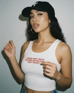 A woman wearing a white ribbed tank top with red text that reads 'WOMEN AREN'T REHAB CENTERS FOR MEN WHO NEED THERAPY', paired with a black baseball cap and denim shorts, posing against a plain light gray wall.