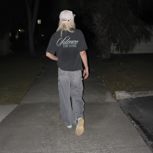 A person walking away on a sidewalk at night wearing a dark gray t-shirt with 'Silence THE NOISE' printed on the back, gray cargo pants, and a white cap.