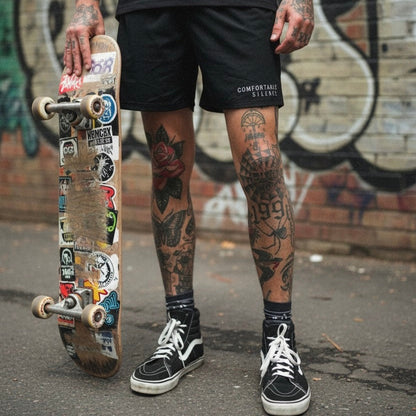 Person holding a skateboard with tattoos visible on arms and legs, standing against a graffiti-covered wall.