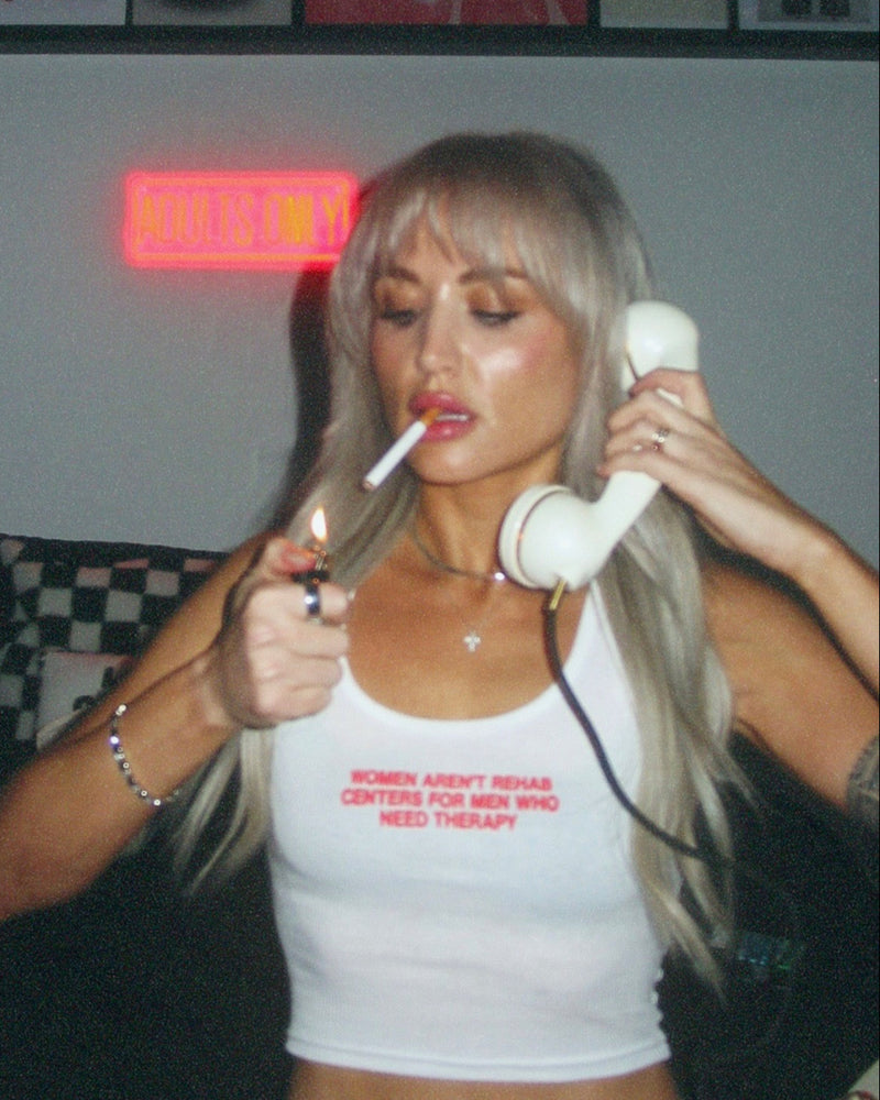 Person holding a lit cigarette and a white phone, wearing a white tank top with text, in a room with posters on the wall.