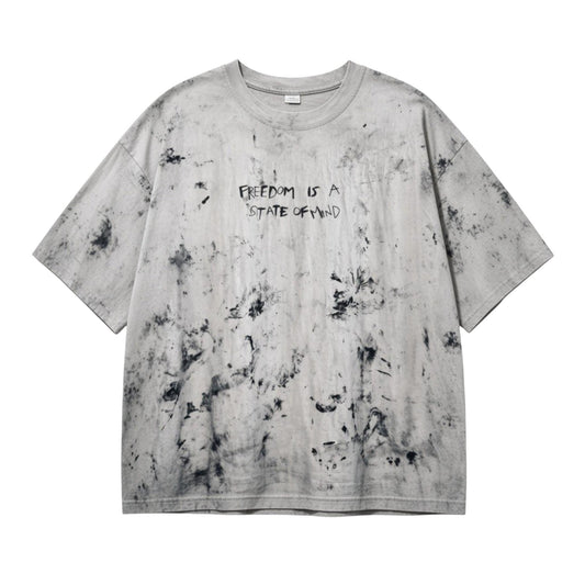 A light gray oversized short-sleeve t-shirt with a black mineral-wash distressed pattern and the text 'FREEDOM IS A STATE OF MIND' printed on the chest, shown flat against a white background.