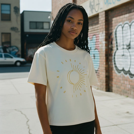 A woman with long black braids stands on a city sidewalk wearing a cream-colored t-shirt with a gold graphic of a sun and crescent moon with the text 'MORNING LIGHT'. The background shows a brick wall with graffiti and a street scene.