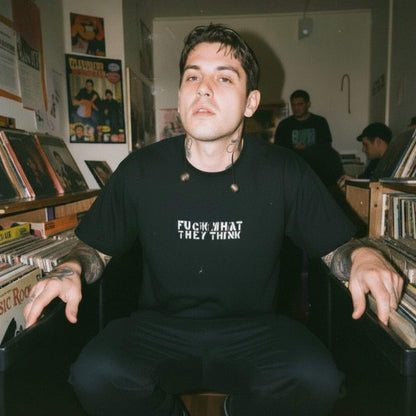 A man sitting in a record store wearing a black t-shirt with the text 'FUCK WHAT THEY THINK' in white. Vinyl records are visible on shelves in the background.