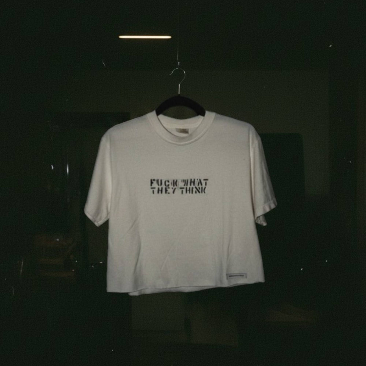 A white short-sleeve t-shirt hanging on a black hanger against a very dark background. The shirt features the text 'FUCK WHAT THEY THINK' printed in black across the chest.