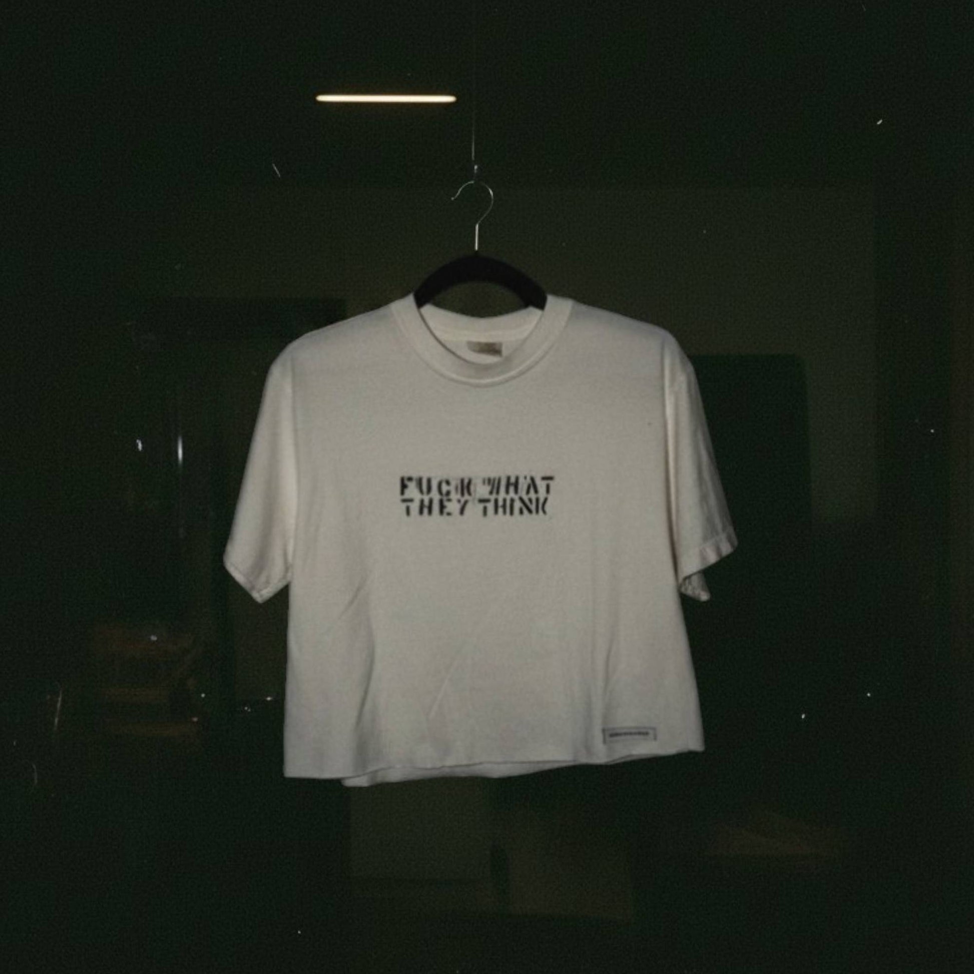 A white short-sleeve t-shirt hanging on a black hanger against a very dark background. The shirt features the text 'FUCK WHAT THEY THINK' printed in black across the chest.