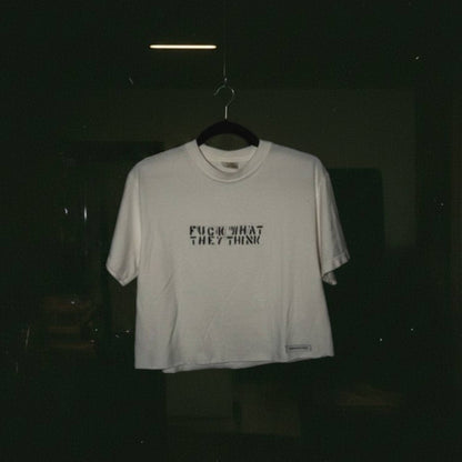 A white short-sleeve t-shirt hanging on a black hanger against a very dark background. The shirt features the text 'FUCK WHAT THEY THINK' printed in black across the chest.