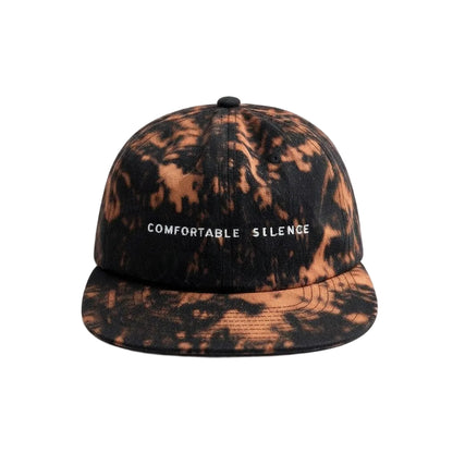 A flat-brim baseball cap with a black and copper-brown tie-dye pattern, featuring the text 'COMFORTABLE SILENCE' embroidered in white on the front.