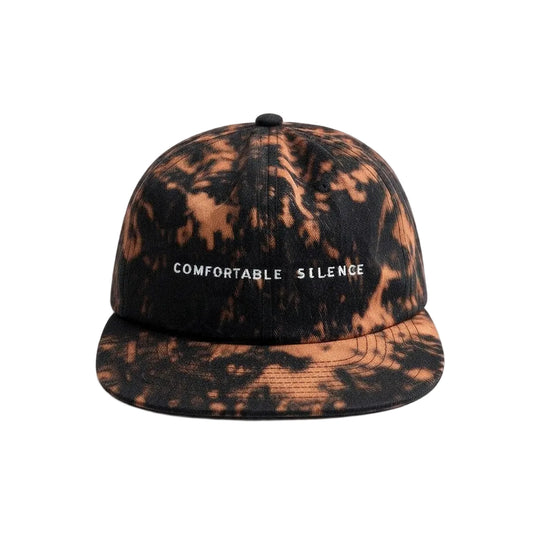 A flat-brim baseball cap with a black and copper-brown tie-dye pattern, featuring the text 'COMFORTABLE SILENCE' embroidered in white on the front.